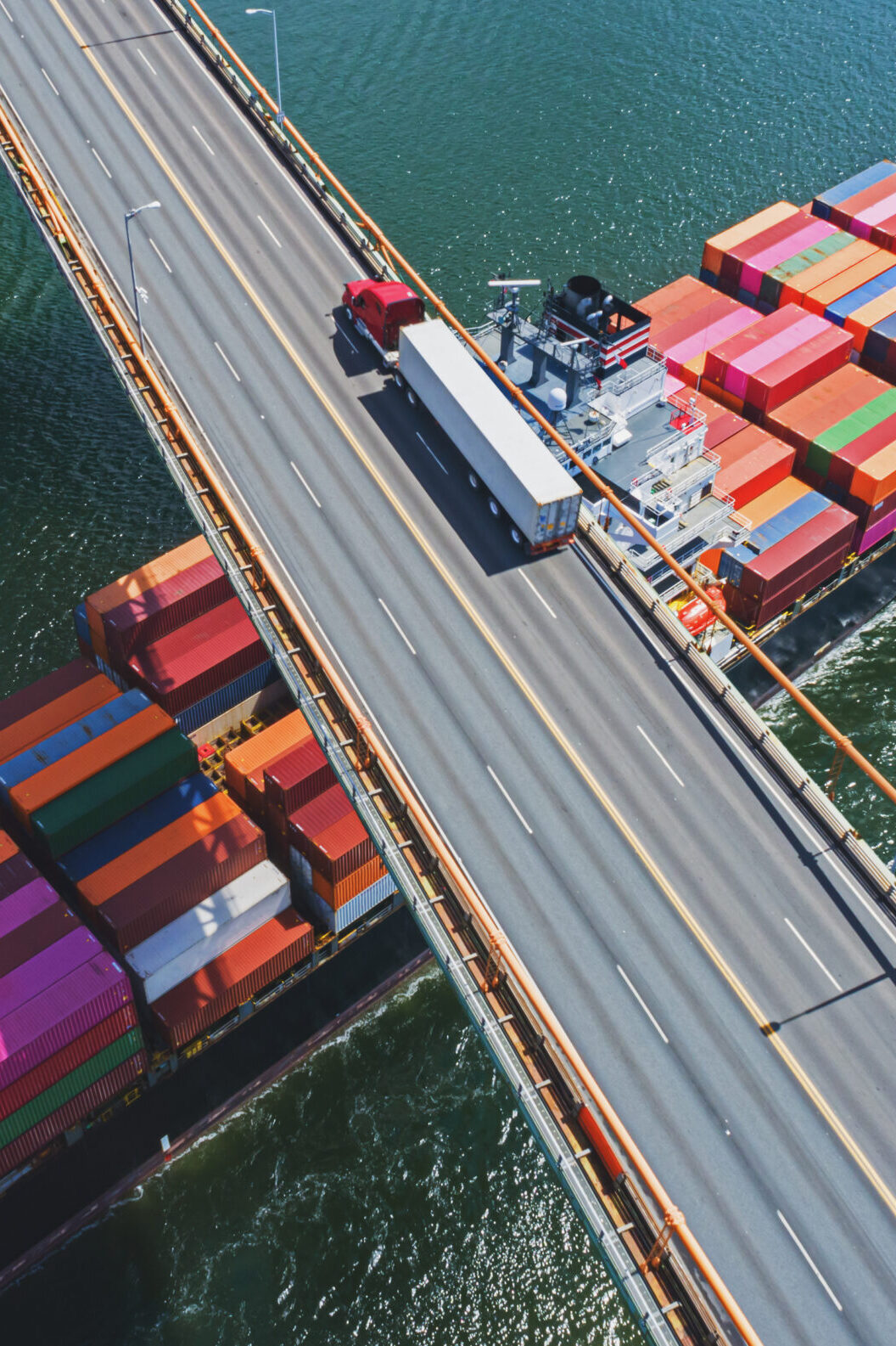 Aerial drone view of a container ship navigating beneath a semi truck crossing a suspension bridge.